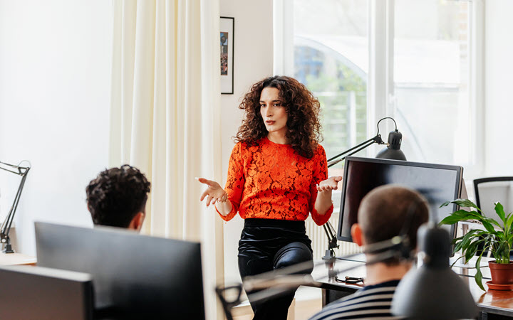 People in an office collaborating. A woman is standing and talking to two men who are seated in front of her.