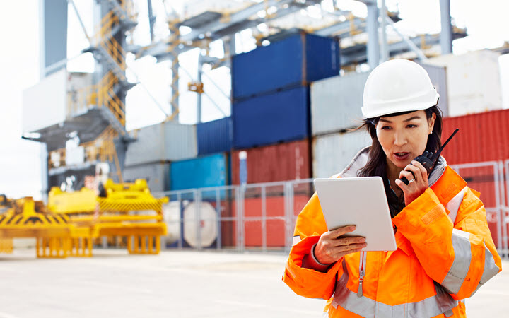 A woman in a construction gear using a walkie talkie and looking at a tablet computer.